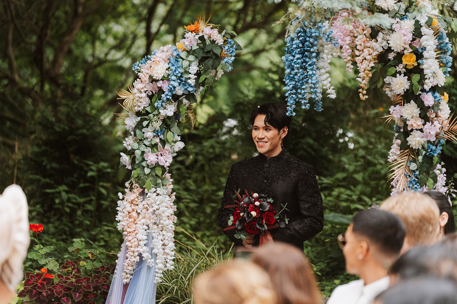 Andrew smiling beneath a floral arch during the ceremony, bouquet in hand